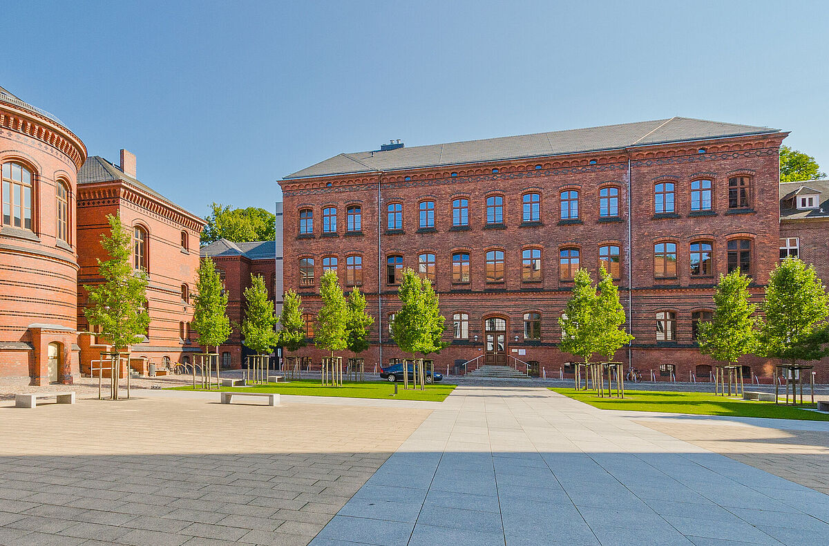 Das neue Verwaltungsgebäude in der Alten Augenklinik Außenansicht der backsteinernen ehemaligen Augenklinik hinter dem begrünten und sonnenbeschienen Innenhof des Historischen Campus vor blauem Himmel.